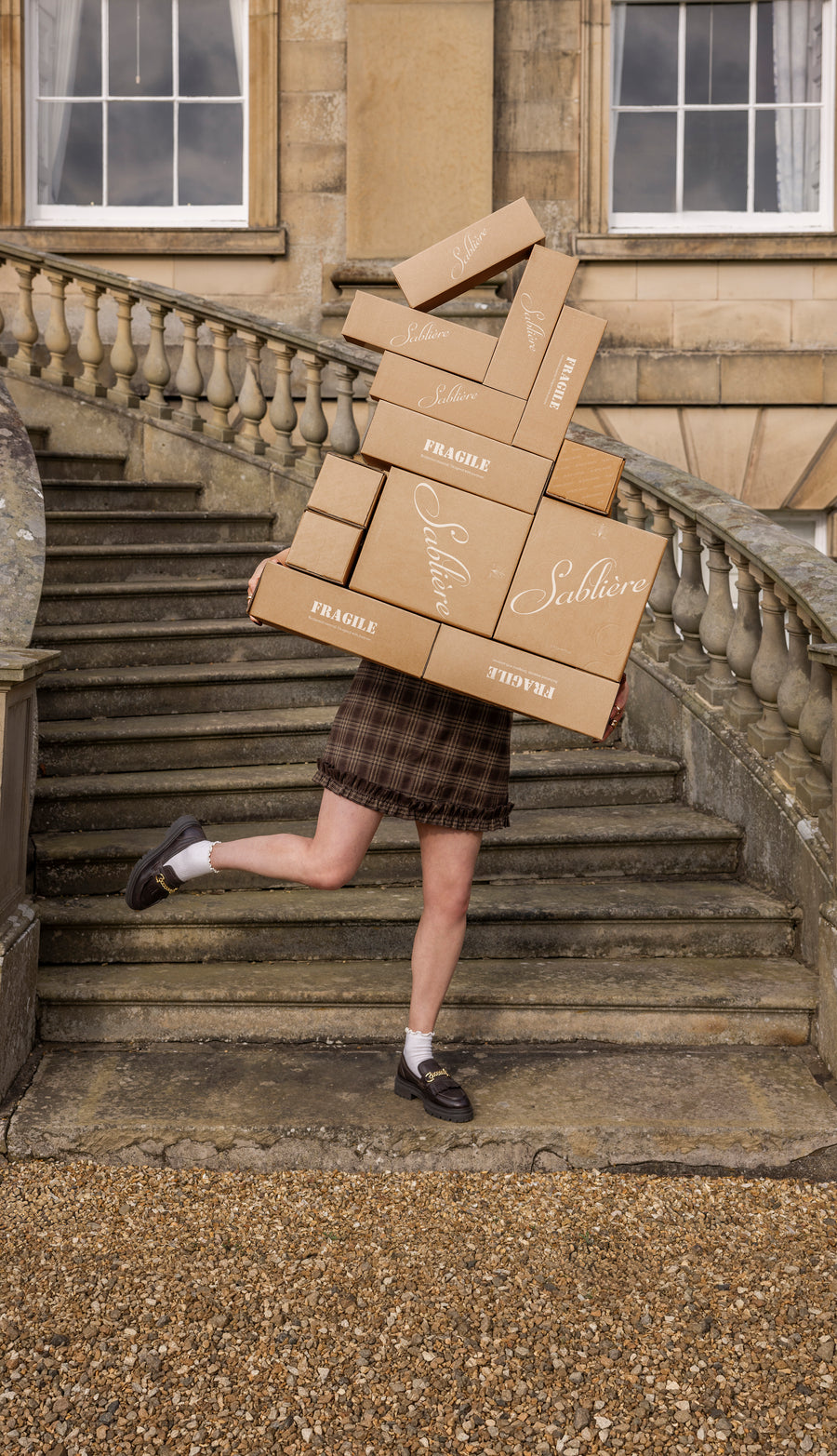 Person carrying stacked boxes labeled Sablière on stone steps with classical architecture in the background