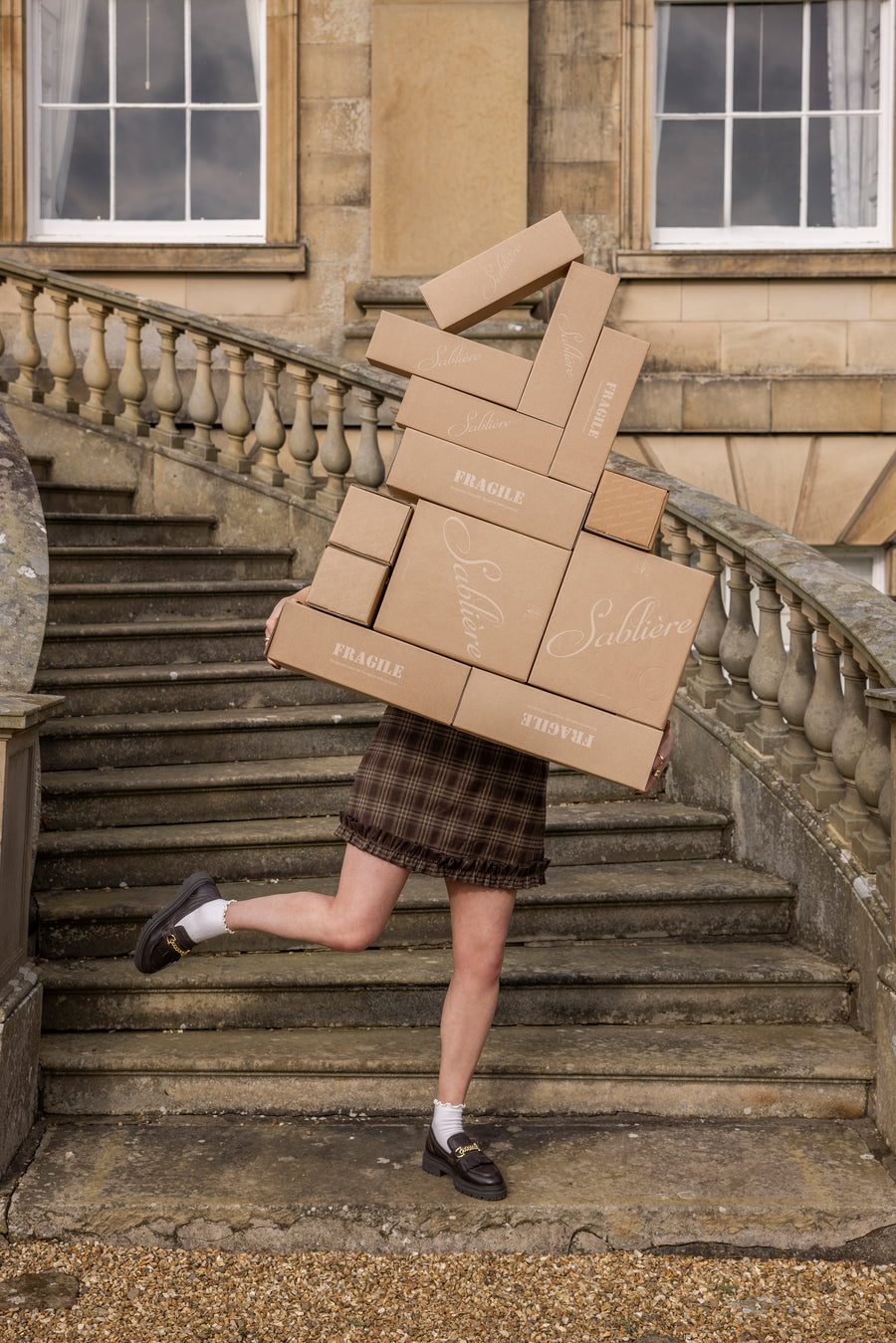 Person carrying a large stack of Sablière boxes on stone steps in front of a building