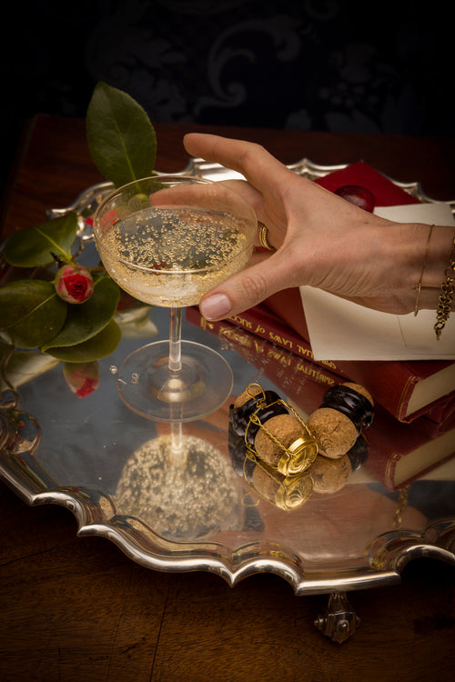 Hand holding a glass of champagne on a reflective surface with books and a bottle in the background.