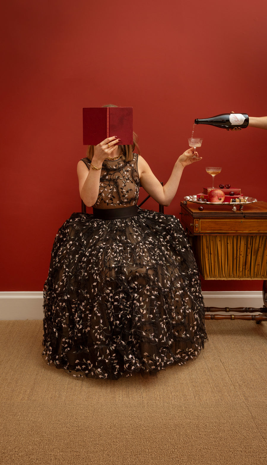 Person sitting in a puffy dress holding a red book and coupe against a red wall with a hand coming in out of shot pouring from a bottle into the coupe